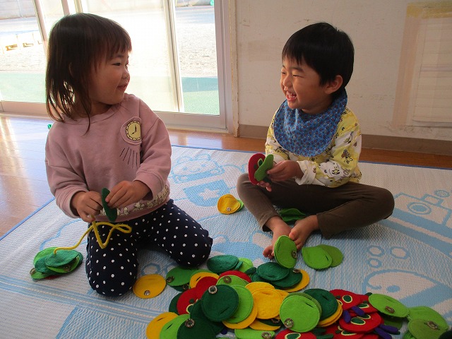 屋内でカラフルなフェルトのおもちゃを床一面に広げ、向かい合って満面の笑みで遊んでいる2人の園児の写真