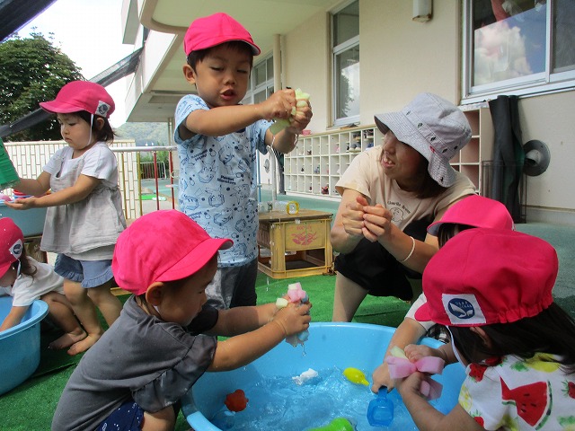 保育教諭とピンクの帽子をかぶった園児たちがたらいに入った水で水鉄砲やスポンジを使って水遊びを楽しんでいる様子の写真