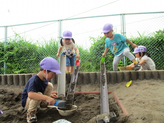 砂場で園児たちが丸太でできた砂場の枠にといを置き、スコップで砂を流して遊んでいる様子の写真