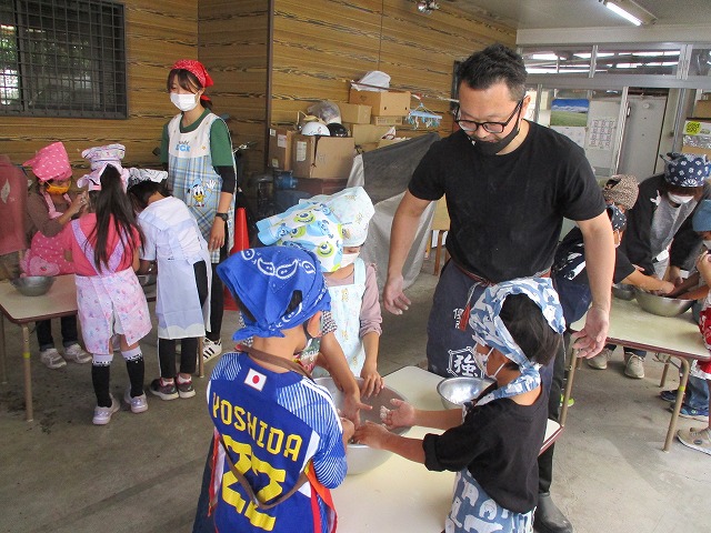 マスクと三角巾をつけた園児たちが、店主に教えてもらいながら真剣な表情でそば打ち体験をしている様子の写真