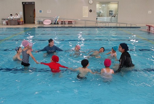 プールで複数の園児たちが保育教諭と手をつなぎ、輪になって楽しそうに遊んでいる様子の写真