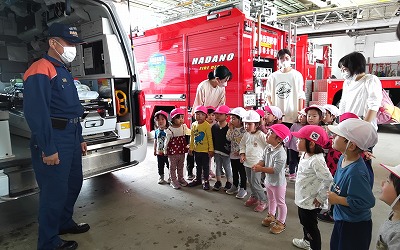 救急車の後ろのドアが開いた状態で消防士が立ち説明をしているのを、消防車の前に並んだ園児たちが熱心に聞いている様子の写真