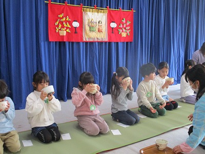 青色の幕にひな祭りの絵のタペストリーが飾られその前で正座をした園児たちが棗を持ち、茶道体験をしている写真