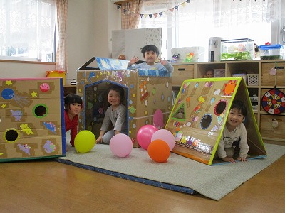 保育園の室内で、段ボールを再利用し、カラフルに装飾された手作りの家やトンネルのような遊具を使って、4人の子どもたちが風船と共に楽しそうに遊んでいる写真