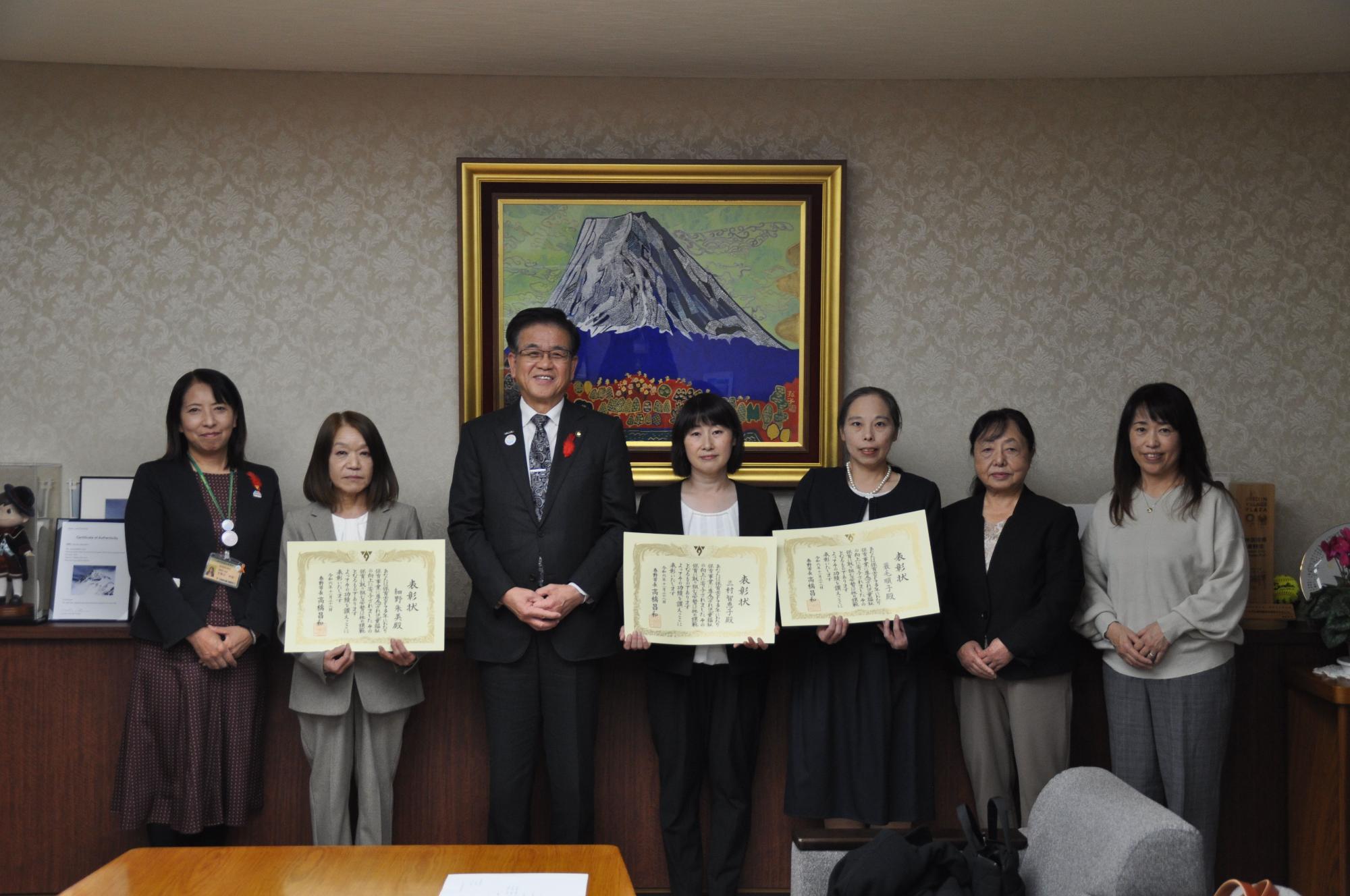 金色の額縁に入った山頂に雪が積もった大きな山の絵画を背景に、市長を中央に保育賞を受賞した3人が賞状を持って立ち、3人の関係者と共に横一列に並んでいる令和6年度表彰式の写真