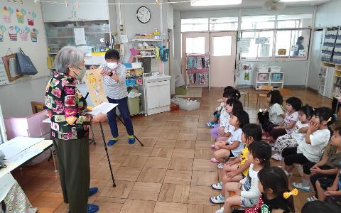 3歳児が並んで椅子に座り、地域の方に紙芝居を読んでもらっている写真