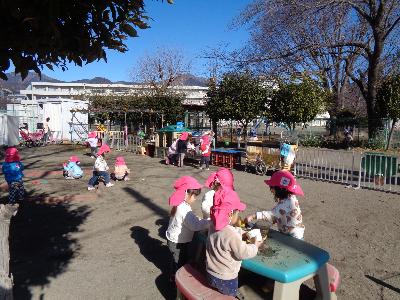 ベンチや家のおもちゃが置かれているこども園の園庭で、ピンク色の帽子をかぶった2歳児がままごとや砂場等で遊んでいる写真