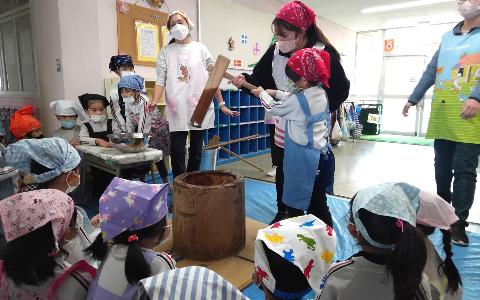 エプロンをつけた5歳児が杵と臼を囲い、順番に保育教諭と一緒にもちつきを体験している写真