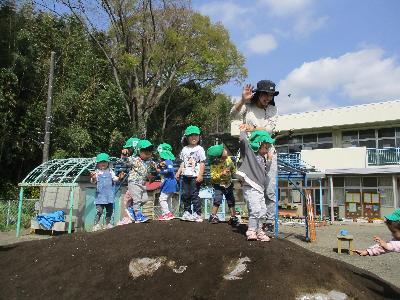 緑色の帽子を被った3歳児と保育教諭が築山の上に登って手を振っている写真
