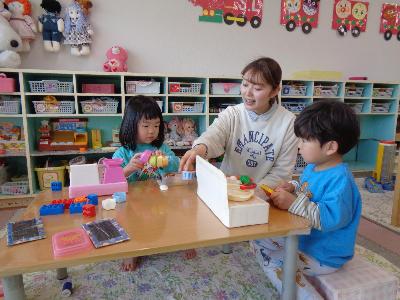 2歳児が保育室でテーブルの上にままごと等のおもちゃを並べて保育教諭と遊んでいる写真