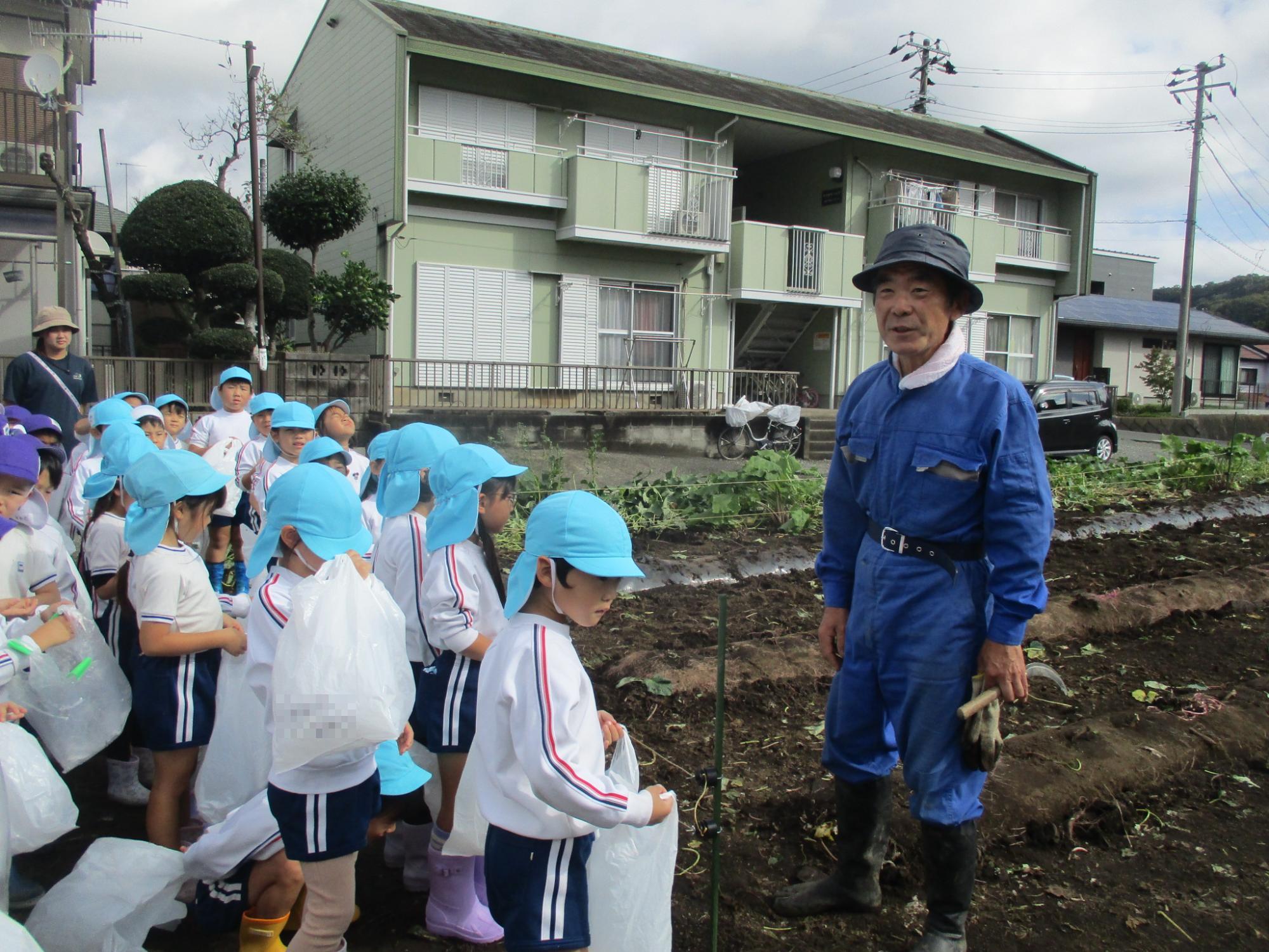 水色の帽子を被った5歳児が地域の方に芋ほりについての話を聞いている写真
