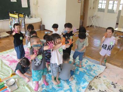 複数の3歳児クラスの子どもたちがピンク色と水色の絵の具を手や足、ローラーを使って大きな紙に塗ったり、手についた絵の具を先生に見せたりしている写真。