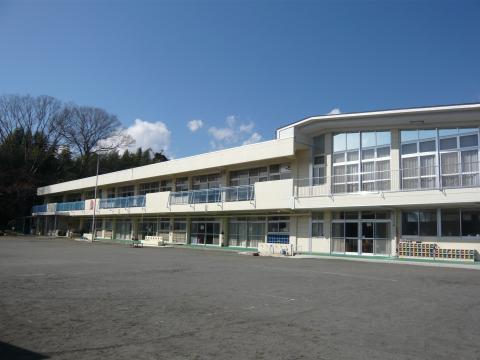 青空の下、周囲には木々が生い茂っているおり、広々とした園庭越しに、白い壁と大きな窓が特徴的なモダンなデザインの二階建ての園舎を撮影した写真