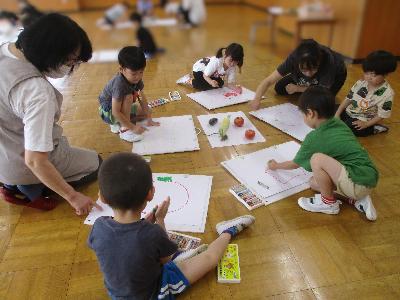 数人の4歳児が絵画指導の先生と一緒に、実際にトマトやトウモロコシを見ながらクレヨンで夏野菜の絵を描いている写真