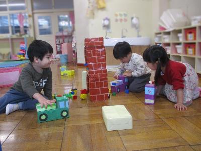 保育室で2歳児の男の子2人と女の子1人が、先生が手作りした車のおもちゃを走らせて遊んでいる写真。