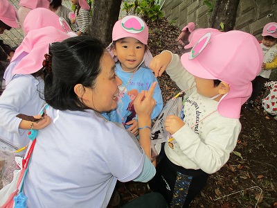 ピンク色の帽子をかぶった園児が、散歩先の小学校で見つけたどんぐりを嬉しそうに保育教諭に見せている様子の写真