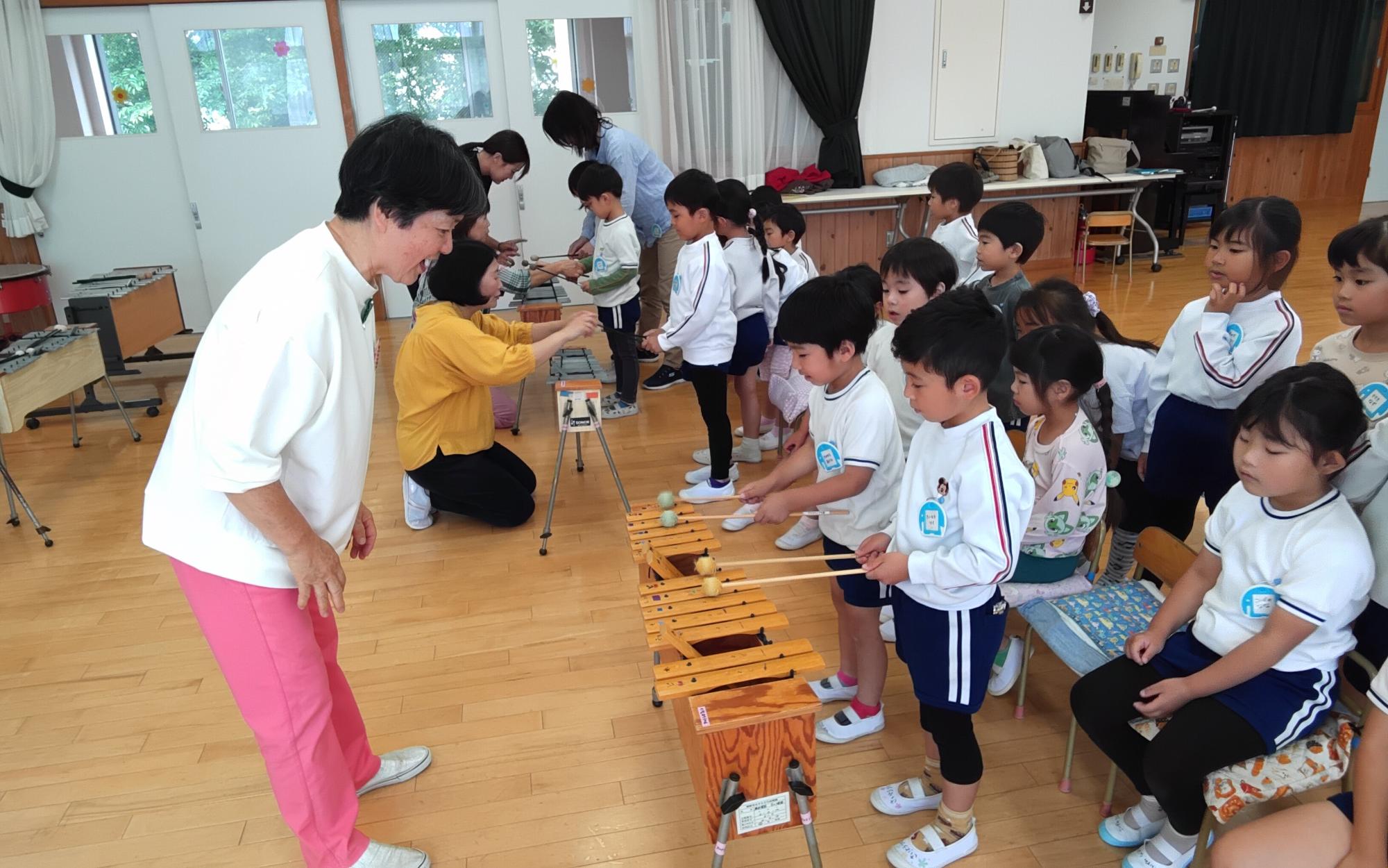 音楽講師に指導を受けながらシロフォンやメタロフォンなどの楽器を鳴らす5歳児の写真