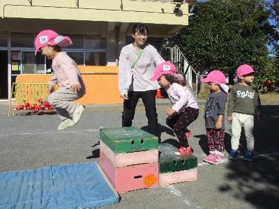 2歳児の男の子一人が先生に見守られ、台からジャンプしておりている。その後ろには楽しそうに順番を待っている子が3名いる写真