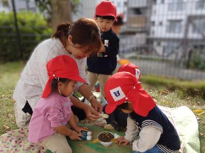 先生と3人の乳児がどんぐりや砂をカップに入れておままごとをしている写真