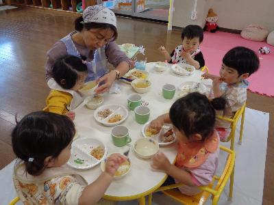 5人の乳児が先生に手伝ってもらいながらも自分で給食を食べている写真。