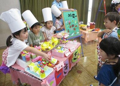 5歳児の園児たちが、ケーキ屋さんごっこをして、年下の友達に廃材で手作りしたケーキを手渡している様子の写真