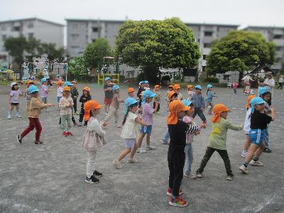 オレンジ色のクラス帽子と水色のクラス帽子をかぶった複数の園児が園庭でリズム体操を楽しんでいる写真
