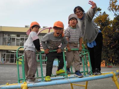 オレンジ色のクラス帽子をかぶった4歳児の園児3名と担任の先生1名がが、ユーホー型のジャングルジムの上に登って微笑んでいる写真