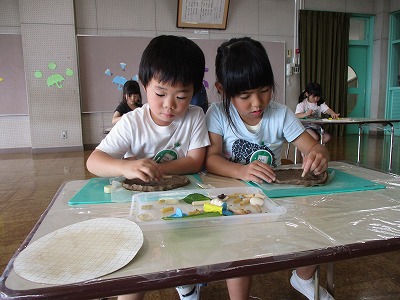 保育室で、二人の園児が粘土に貝殻や葉っぱなどの自然素材を使用して模様付けをして、夢中でお皿作りをしている様子の写真