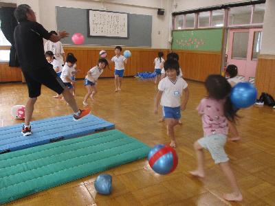 園の遊戯室で、複数の園児と体操の先生がボールを投げ合って遊んでいる写真