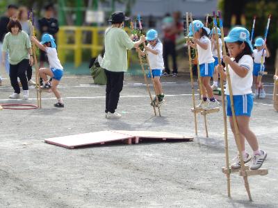 水色のクラス帽子をかぶった5歳児数名が園庭で竹馬に乗っている写真