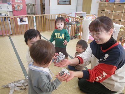 保育室で、保育教諭が破いて丸めた新聞紙を0歳児の園児から笑顔で受け取っている。周りの3人の園児はその姿を興味をもって見ている様子の写真