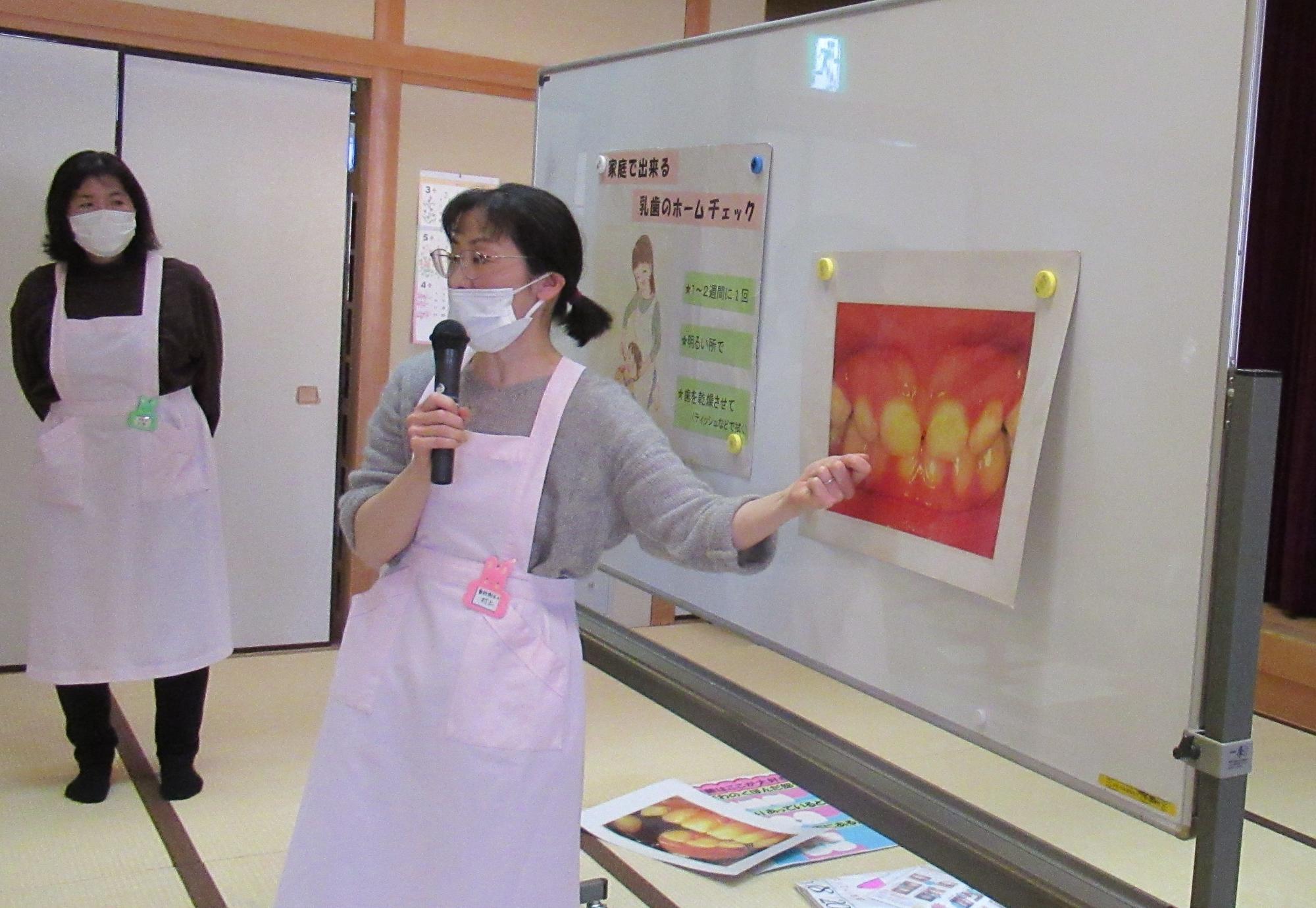 エプロンとマスクを着用した女性が室内でマイクを持ち、ホワイトボードに貼られた歯の黄ばみと歯茎の炎症を示す写真を指さしながら説明している幼児食と歯のセミナーの写真
