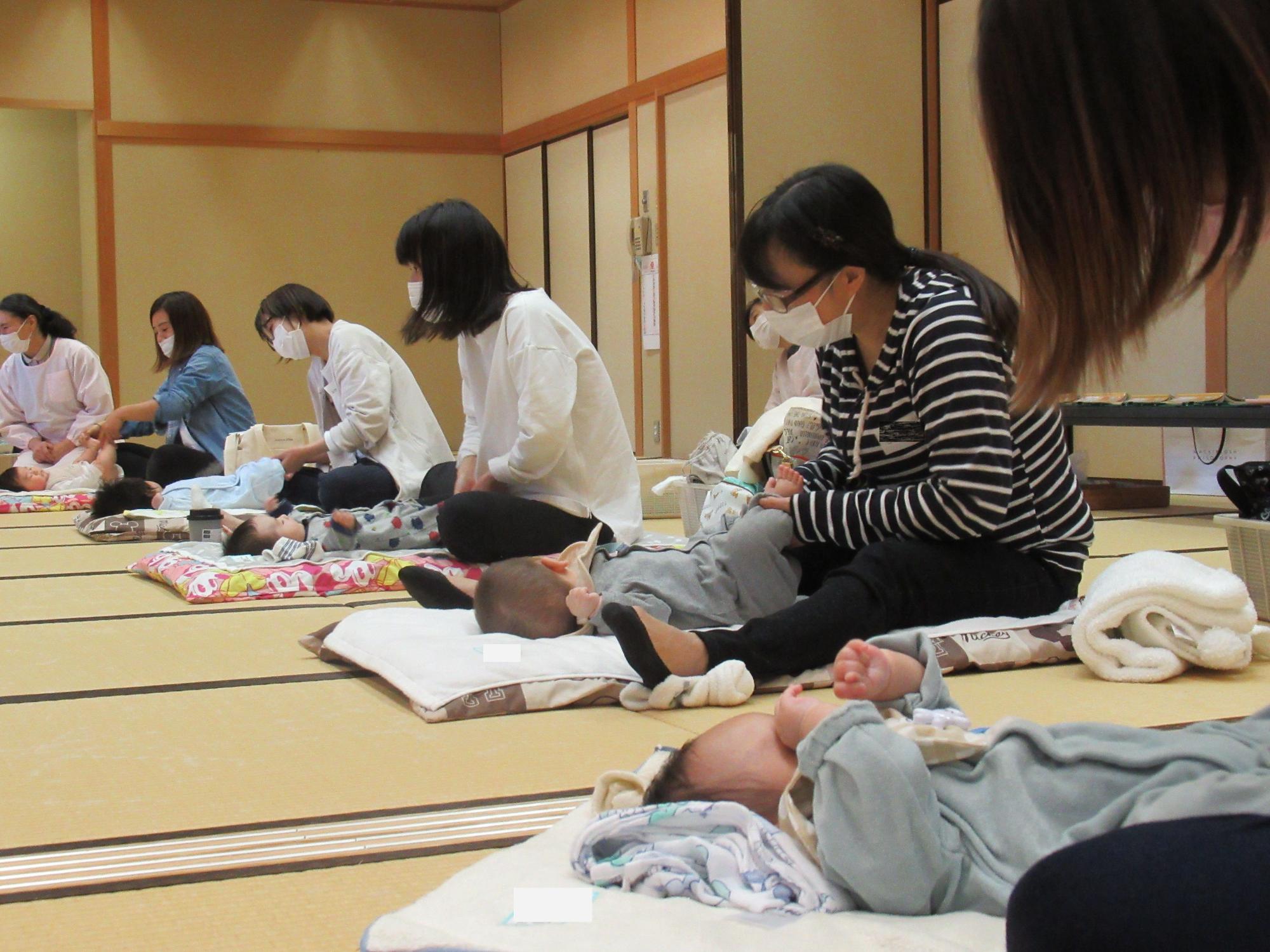 和室に敷かれた布団の上に仰向けに寝かせられた赤ちゃんの横で、マスクを着用した母親たちが座ってベビーマッサージを行っている様子の写真
