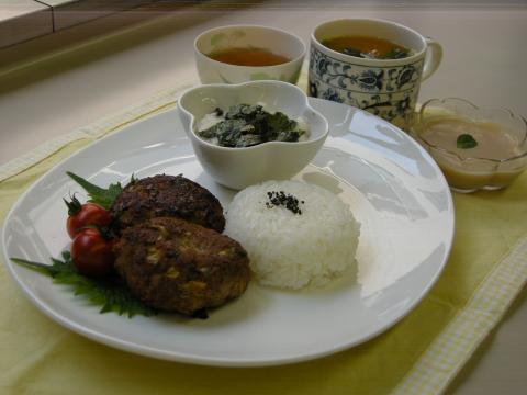 平らな白い丸皿に、大葉の上にミニトマト2個と丸いハンバーグ2個、黒ごまが振られた丸いご飯、海苔が乗った和え物が盛られ、スープとデザートが載った写真