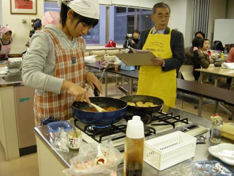 エプロンと白いバンダナを身につけた女性が、コンロの上でフライパンの中の料理を木べらでかき混ぜ、その隣で黄色のエプロンをつけた男性がクリップボードを持って調理の様子を観察している様子の写真