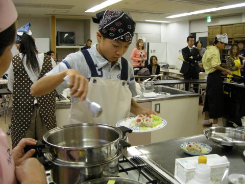 バンダナとエプロンを着用した男性が、調理台でステンレス製の寸胴鍋から盛り付けられた皿へ料理をよそっている様子の写真