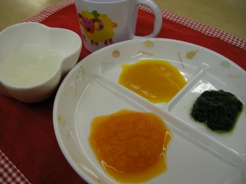 黄色と緑色のペースト状の食材とオレンジ色のピューレが三つに仕切られた白いプレートに盛られ、隣には白いお皿に入った液体と羊の絵柄のマグカップが置かれている離乳食の写真
