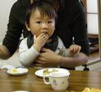大人の膝の上に座ってテーブルで食事をしている子供が、手づかみ食べする様子の写真