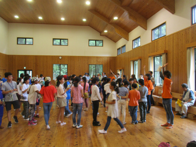 木造の広々とした室内で、オレンジ色のTシャツを着たスタッフが、大勢の子供達にレクレーションの動きを教えている写真