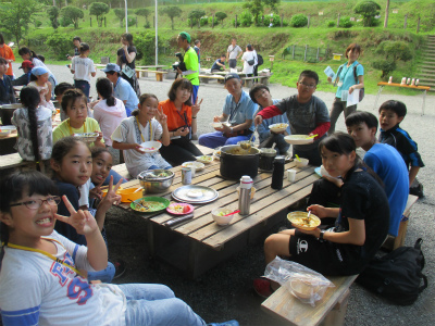 屋外に設置された木製のテーブルを囲み、子供達と大人がカレーを食べながら、ポーズをとっている写真