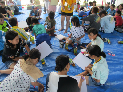 ブルーシートの上で、子供達が輪になって座り、色紙にペンでメッセージを書いている写真
