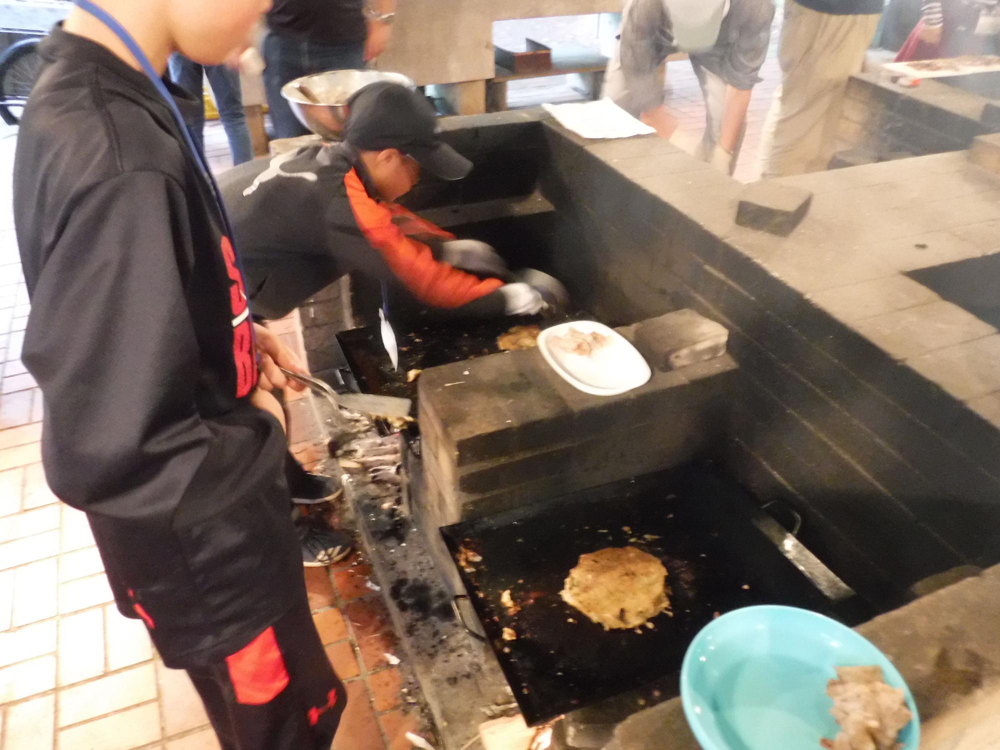 屋外の調理場で、子供達が鉄板の上でお好み焼きを焼いている写真