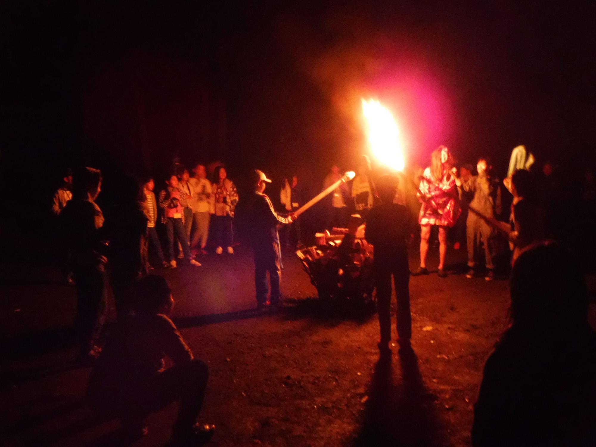 暗闇の中、燃え盛る大きな炎を囲み、多くの子供達や大人が見守りながらキャンプファイヤーを楽しんでいる写真