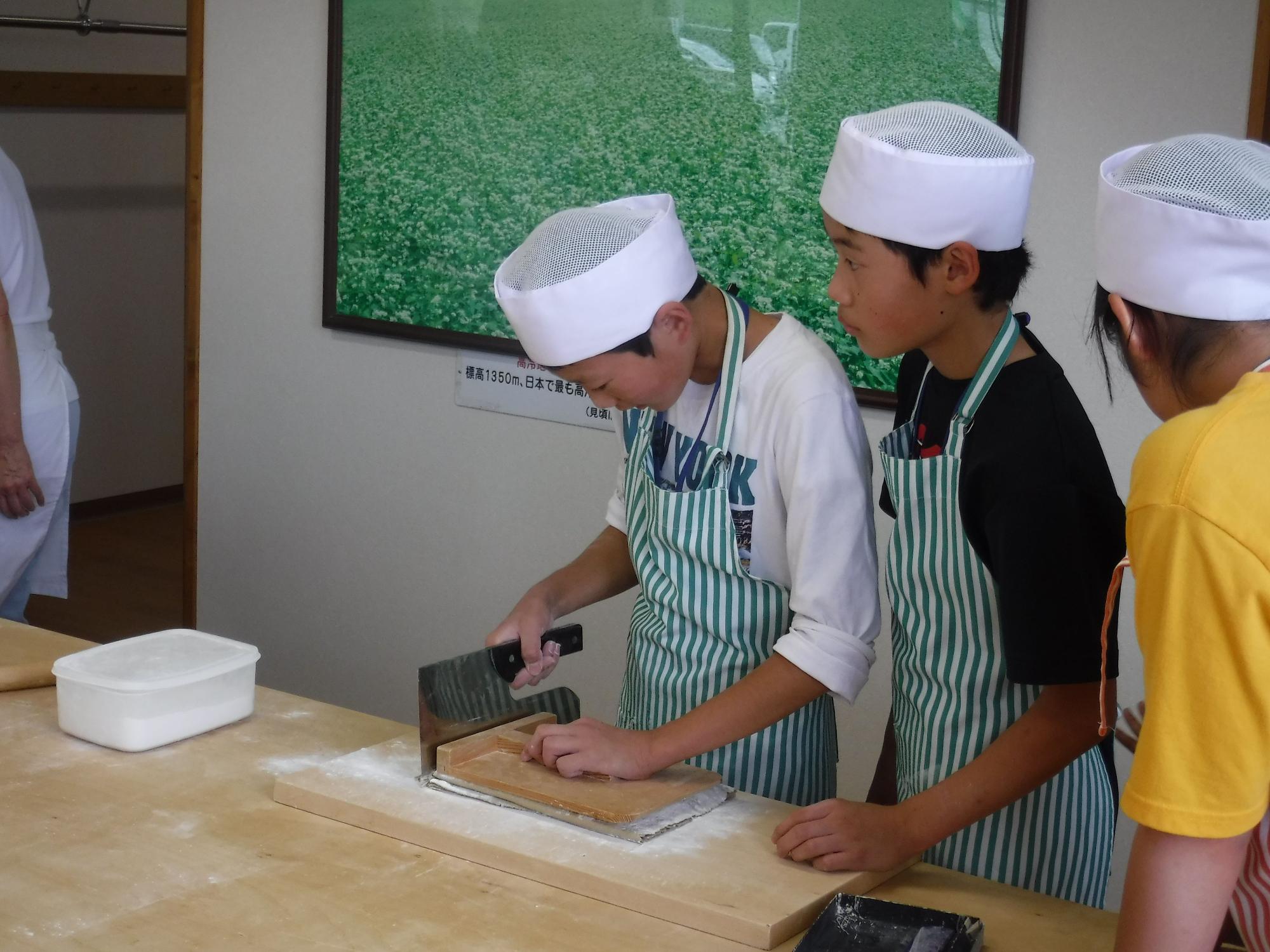 エプロンに白い帽子をかぶった子供達が、そば打ち台の上でそばを切っていたり、作業を眺めているいる写真