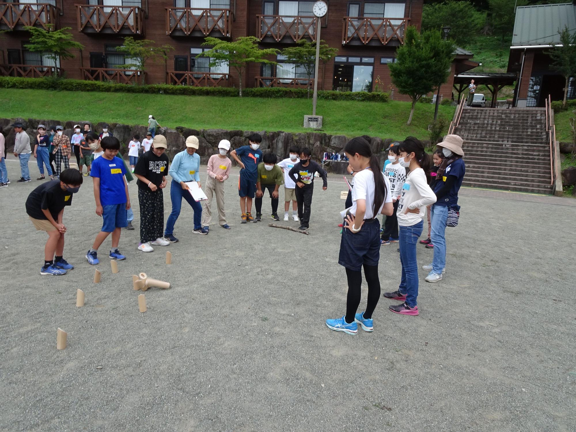 表丹沢野外活動センターの広場で、木製の棒を使って木製のピンを倒している子供と、その様子を楽しそうに眺めている子供達の写真