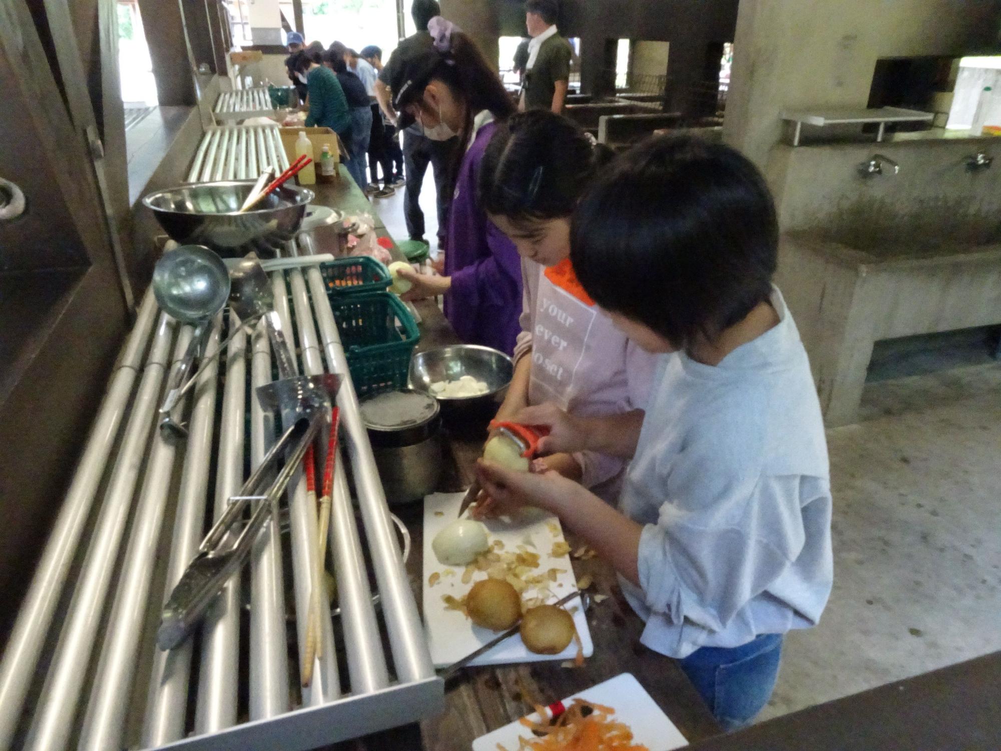 厨房で、子供達が玉ねぎの皮を剥いたり食材をカットしたり、野外炊飯の準備に取り組んでいる様子の写真