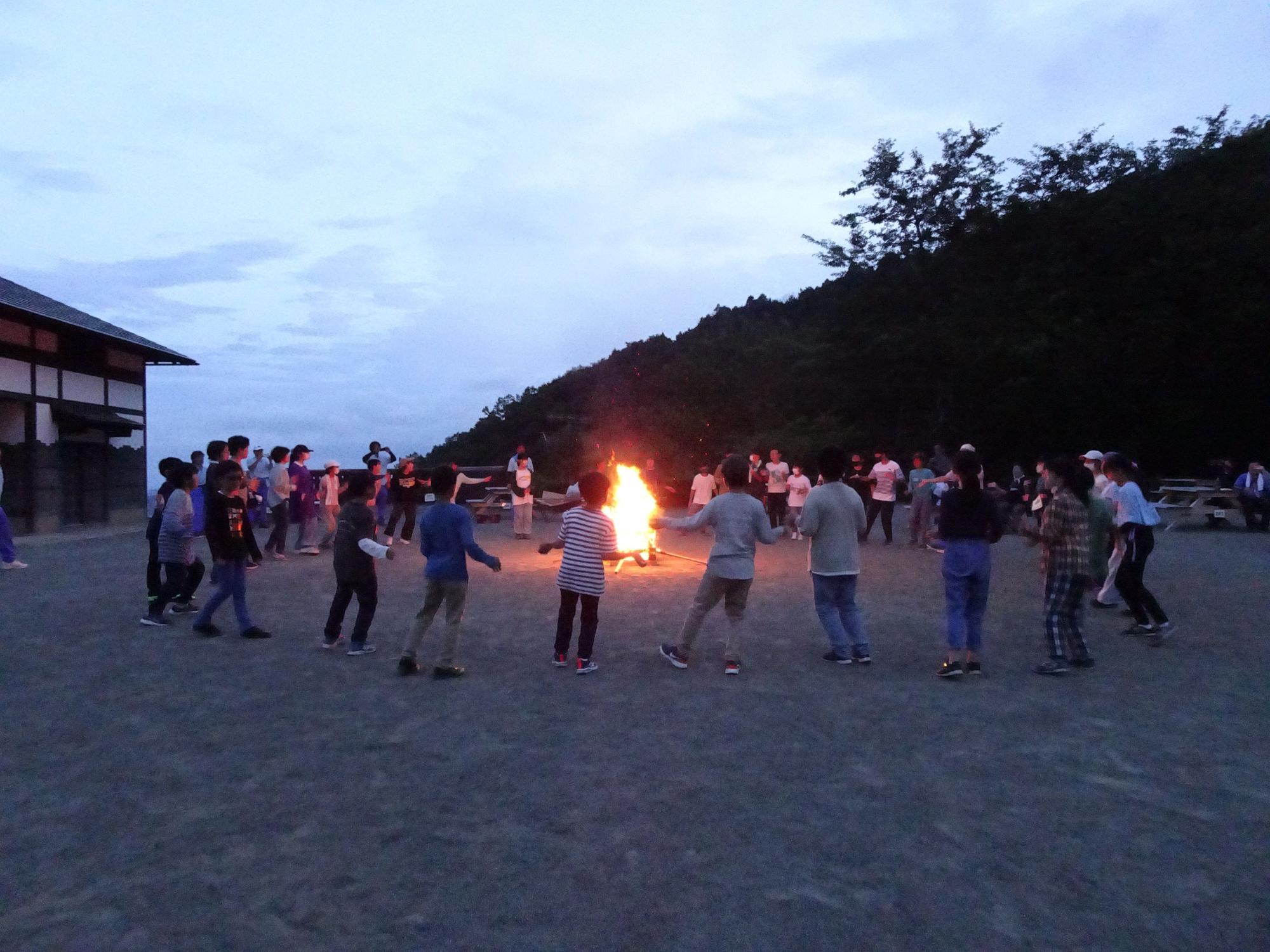 薄暗い広場で、たき火の周りに子供達が集まり、キャンプファイヤーを楽しんでいる写真