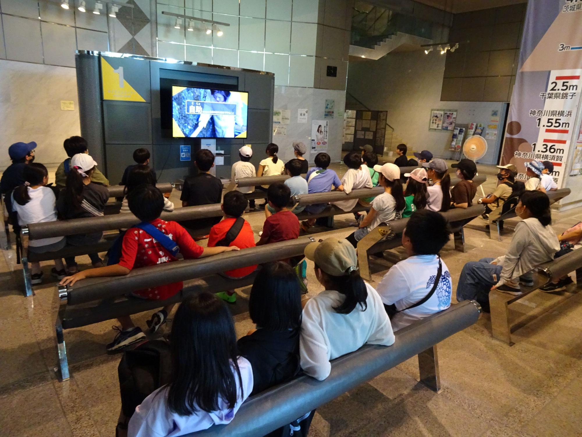 広い室内のベンチに大勢の子供たちが座り、正面のモニターに映し出された映像を鑑賞している写真