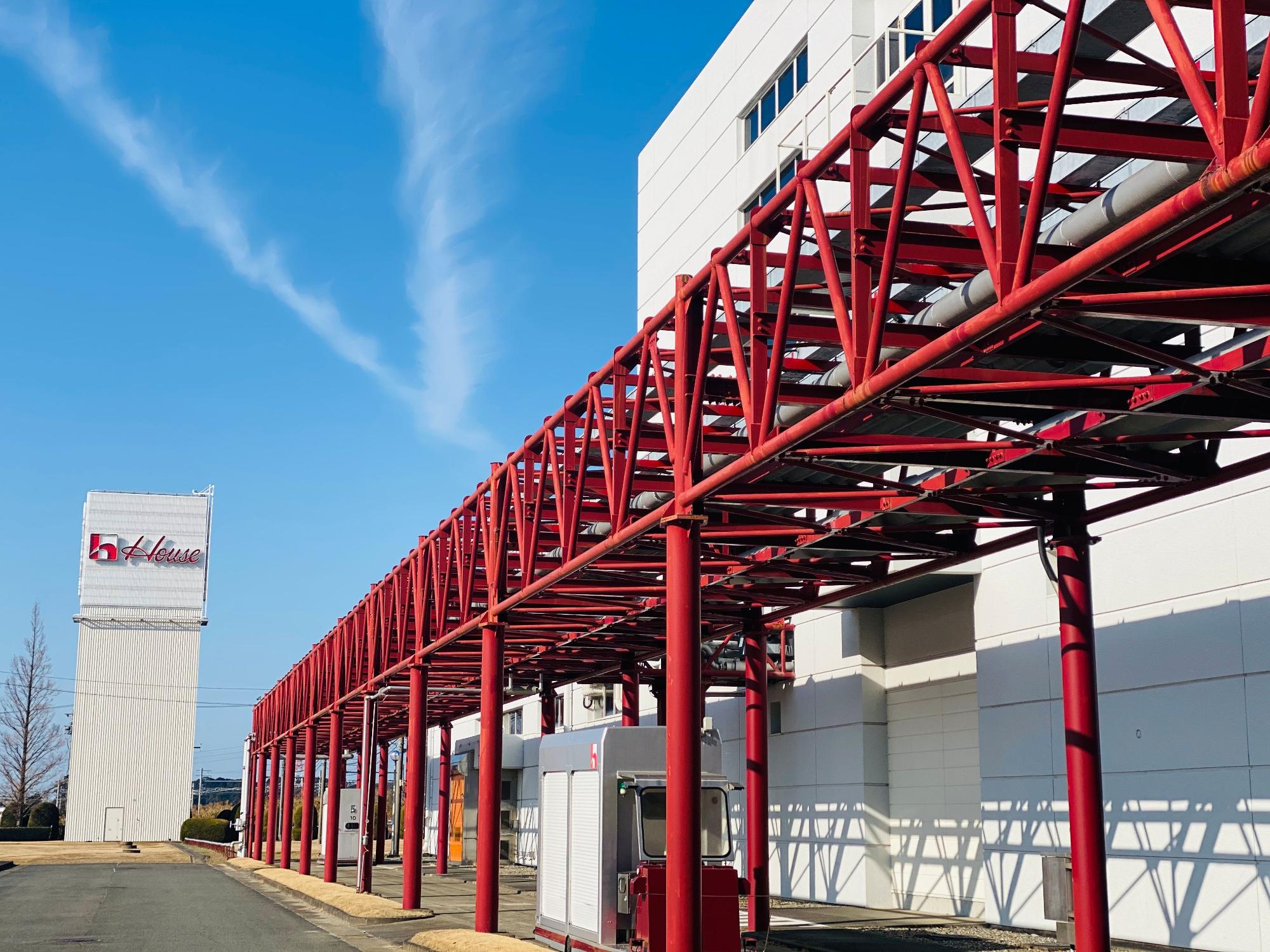 青空の下、白い建物に沿って赤く塗られた複雑な鉄骨構造の連絡通路が伸びており、奥には、「House」のロゴが掲げられた白い看板が立っている写真