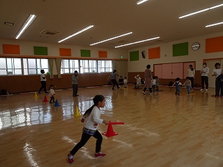 広い室内で、子供がバトンを持ちコーンの周りを駆け回ったり、大人と子供達が体操をしている写真